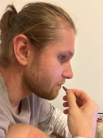 A close-up shot depicts a light-skinned person with blond hair pulled back in a low bun and a reddish-brown beard. The person is in profile, looking upwards. A hand is holding a dark-colored object near the person's mouth. The person is wearing a gray t-shirt with a white long-sleeved shirt underneath. The location is not discernible from the image.