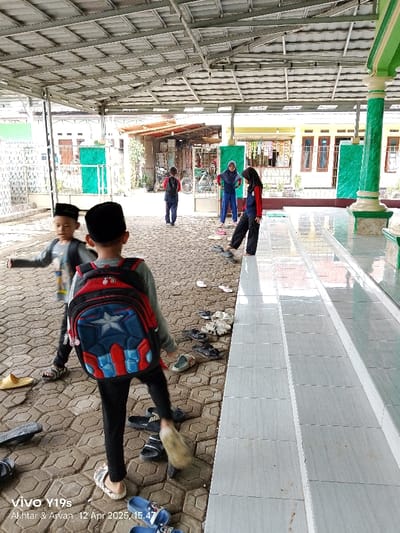 The image shows a group of children in Anjatan, Indonesia, located at coordinates -6.419 latitude and 107.979 longitude. The children are all wearing school uniforms and are standing in front of a building. The building appears to be a mosque. The image is taken from the perspective of a person standing on the steps leading up to the building. There are a few sets of shoes left on the steps. Some children are wearing backpacks. There is a green column behind the children.  The area appears to be an outdoor courtyard with a concrete floor and a metal roof.  The children are interacting with each other and appear to be chatting.  The environment is relatively quiet, and the children are in a relaxed and friendly atmosphere.