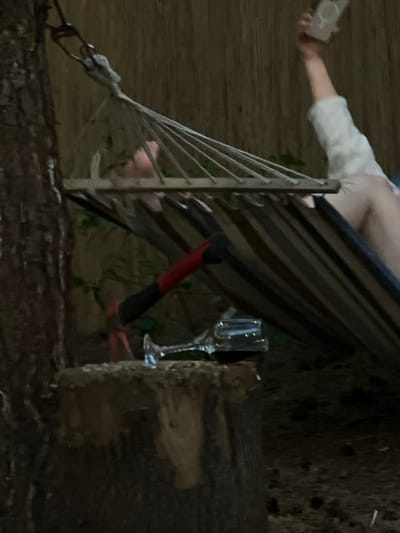 In a dimly lit outdoor setting, possibly a backyard or campsite, a hammock is suspended between a tree and an unseen anchor point. A person, only partially visible and lying in the hammock, is holding up a mobile phone, suggesting they are taking a selfie or recording a video. Below the hammock, an axe with a red and black handle rests on a tree stump, positioned near a wine glass. The environment appears natural, with a wooden fence or screen in the background. The scene is suggestive of a relaxed, perhaps slightly unconventional, moment outdoors.