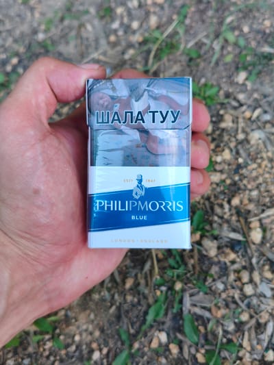 A hand holds a white cigarette pack with blue and white branding. The top of the pack features Cyrillic text in white, "ШАЛА ТУУ," above an image of what appears to be a person's face. Below this, the brand name "PHILIP MORRIS" is displayed prominently in white text on a blue band. Beneath that, the word "BLUE" is written in smaller white text. At the bottom of the pack, "LONDON ENGLAND" is printed in small gold letters. The background consists of dry ground with scattered pebbles and some green vegetation. The image was taken outdoors, likely in Zerendi, Kazakhstan, given the presence of Cyrillic script on the cigarette pack, which is commonly used in countries that were part of the former Soviet Union, including Kazakhstan.