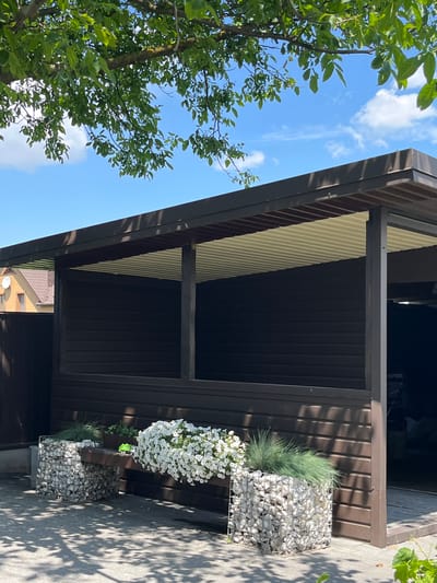 A low-angle shot captures the side of a dark brown, slatted building, likely a house or shed, with a covered porch area. The roof of the porch is a light yellow corrugated material, supported by dark brown beams. To the left of the building, a large tree with vibrant green leaves dominates the frame, reaching up towards a bright blue sky with scattered white clouds. In the foreground, on the right side of the image, a gabion basket filled with white stones sits next to a smaller gabion basket containing lush green grass and a cluster of white flowers. The ground appears to be paved with light gray stones, and dappled sunlight creates shadows on the ground and the side of the building. The image conveys a sense of a warm, sunny day, likely in the late morning or early afternoon, with pleasant weather. The overall impression is of a well-maintained outdoor space in a residential setting.