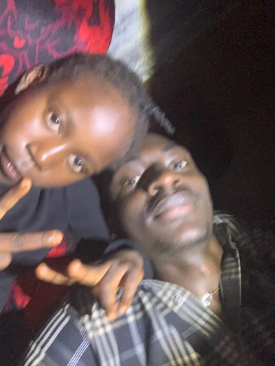 The image shows a close-up selfie of two young Black individuals. The person in the foreground is looking directly at the camera with their mouth slightly open, revealing teeth. Their right hand is raised, with two fingers extended, forming a peace sign. A second person is lying down behind them, with their head turned away from the camera, looking upwards. This person is wearing a plaid shirt with a silver necklace visible. The background is dark and indistinct, suggesting an indoor setting or nighttime. The overall mood appears casual and friendly. The image was taken in Russo, Nigeria. There is no visible text in the image.