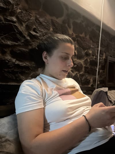 The image, rotated 90 degrees clockwise, features a young person lying on their side against a dark, textured stone or brick wall, typical of an old building in Belopoptsi, Bulgaria. The dim lighting suggests it is evening or night indoors.
The subject has dark, slicked-back hair and a neutral expression, looking upwards. They are wearing a white t-shirt, with a glimpse of a light pink garment or design visible beneath it on their right shoulder and chest. A black string bracelet is on their left wrist. Their hands are clasped, holding a smartphone, which is partially visible in the upper right.
Faint, indistinct text, possibly a tattoo like "Je te veux", can be seen on their right inner arm. A thin white wire or string crosses horizontally through the upper background. The stone wall provides a rustic backdrop, and a dark, rectangular shape (possibly a window or frame) is visible higher on the wall. The person's right arm rests on a light grey, textured surface, possibly a cushion or bedding. The activity appears to be the person relaxing and engaging with their phone.