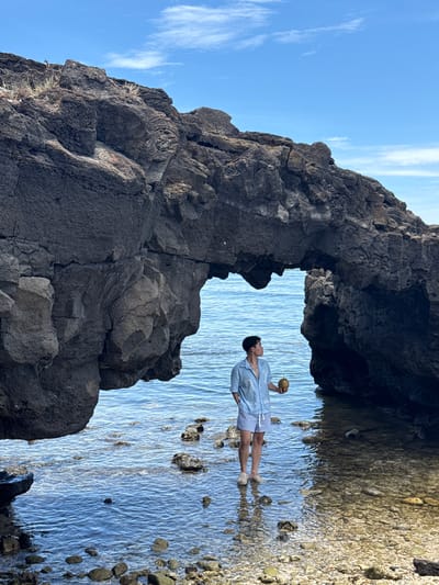 A person stands in shallow ocean water near a rocky arch formation. The person, facing away from the viewer and to the right, is holding a coconut. They are wearing a light blue short-sleeved collared shirt with a white graphic design and blue and white striped shorts, and light-colored shoes. The water is clear and shallow, with rocks and pebbles visible on the seafloor. The rocky arch, dark gray in color and irregular in shape, frames the ocean view. The sky above is blue with scattered white clouds. The scene appears to be on Lý Sơn Island, Quảng Ngãi Province, Vietnam.