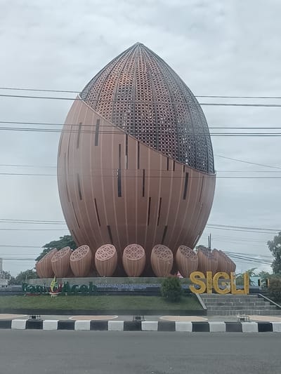 This image depicts the Sigli welcome gate, a prominent landmark in the city of Sigli, Indonesia. The main subject is a large, egg-shaped sculpture with a textured, patterned top section and a smooth, segmented lower section. The sculpture is situated in a roundabout, with a road in the foreground.
To the left of the sculpture, the words "Bank Aceh" are visible in green lettering, along with a stylized logo. To the right, the word "SIGLI" is spelled out in large, yellow 3D letters on a tiered stone structure. Surrounding the base of the sculpture are several smaller, segmented objects that resemble halves of the larger sculpture, arranged in a semi-circle.
The area around the sculpture is landscaped with grass and some bushes. In the background, trees and buildings are visible, along with a cloudy sky. Several power lines are strung across the upper portion of the image, obscuring some of the sky and the top of the sculpture. The overall impression is of an outdoor public space with a focus on civic identity and branding.