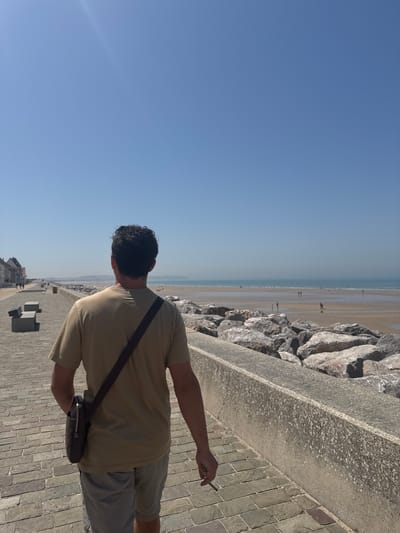 A person stands on a paved walkway overlooking a beach with the ocean in the background. The person is seen from behind, wearing a tan t-shirt and gray shorts. They have dark, curly hair and are carrying a dark bag with a strap across their chest. In their right hand, they hold what appears to be a cigarette. The beach is populated with a few distant figures.

The scene is bathed in bright sunlight, suggesting a clear day. The walkway is made of brick pavers, and to the left of the walkway is a low wall constructed of large, rough stones. Beyond the stones, the beach stretches out towards the water, with the ocean meeting the horizon under a vast, clear blue sky. In the far distance, along the coastline, there are some buildings and structures visible. The overall atmosphere appears relaxed and casual, typical of a sunny day at the seaside. The image was taken in Wissant, France. There is no visible text in the image.