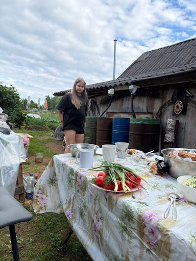 The image depicts an outdoor setting in Bronnitsy, Russia, featuring a young girl standing near a table laden with food. The table is covered with a floral tablecloth and set with various dishes, including a bowl of tomatoes and green onions, a large metal bowl containing bread and possibly meat, and other serving dishes. Two cups and a white mug are also visible on the table. In the background, there are several large barrels and a rustic wooden structure. The ground is covered in grass, and there is evidence of a garden or planted area. The sky is overcast with clouds. The girl is wearing a black t-shirt and shorts. The overall scene suggests a meal is being prepared or has recently taken place outdoors.
