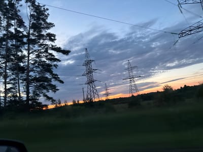 A serene sunset illuminates the sky over a rural landscape in Garevaya, Russia. Tall pine trees stand silhouetted against the dusky sky on the left, their branches reaching towards the clouds. In the middle ground, imposing power line towers stretch upwards, their metallic structures stark against the colorful horizon. The sky transitions from a pale blue at the top to soft orange and yellow hues near the horizon, with dramatic, wispy clouds scattered throughout. A dense line of trees and vegetation forms a dark band across the landscape, obscuring the lower parts of the power lines and creating a sense of depth. The foreground is dominated by blurred green foliage, suggesting the image was taken from a moving vehicle. The overall impression is one of peaceful, natural beauty juxtaposed with human infrastructure.
