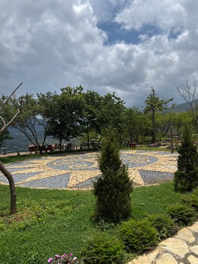 The image shows a landscaped area in Sa Pa Ward, Vietnam. A stone mosaic path with a circular design is flanked by green grass and shrubbery. Trees line the path, and wooden tables and chairs are visible underneath the trees. In the background, a mountain range is partially obscured by a cloudy sky with patches of blue.