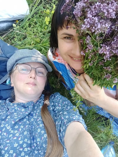 Two women are lying on their backs on the grass. The woman on the left is wearing a blue floral dress and a grey checkered cap. She has glasses and her long blonde hair is braided. The woman on the right is holding a bouquet of small purple flowers in front of her face and is wearing a colorful striped shirt. The background is grassy with some green plants and yellow leaves. A backpack is visible to the left of the woman in the cap. The image was taken in Shpaniv, Ukraine.