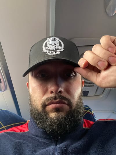 A close-up selfie features a man with a beard and a black trucker hat. The hat has a white embroidered logo depicting a skull with a barbell and text. The man is wearing a navy blue fleece jacket with red and yellow accents. He is holding the brim of his hat with his right hand, and his face is illuminated from the front and slightly from above. The background appears to be the interior of a vehicle, possibly a bus or train, with seats visible behind him. The lighting suggests an indoor environment. The image does not provide any information about the location in the city of Koskolovo, Russia.