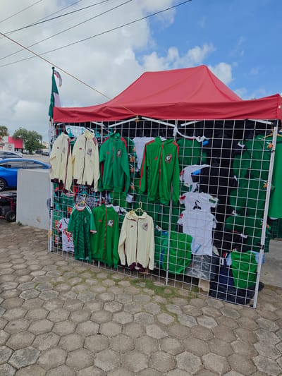 A display of merchandise, likely jerseys and jackets, is set up outdoors under a red canopy tent. The items are predominantly green and white, with some yellow jackets also visible. They appear to be sports apparel, possibly related to a soccer team, given the presence of what looks like the Mexican flag flying from a pole to the left of the tent. The merchandise is hung on a white wire grid. In the background, a blue car is parked next to a grey concrete structure. The ground is paved with hexagonal stones. The sky is partly cloudy with blue patches. The overall scene suggests a casual outdoor market or roadside vendor in Cancún, Mexico, likely during the daytime. No people are directly visible in the image, but the setup implies a commercial activity. There is some text visible on some of the clothing, including what appears to be a logo with "Mexico" on it.