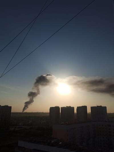 The image depicts a cityscape in what appears to be Кондратово, Russia. Silhouetted apartment buildings line the right side of the frame. In the distance, dark smoke rises from the ground, obscuring the sun and creating a haze in the sky. Electrical wires stretch across the lower left of the image. The sky transitions from dark blue on the left to a hazy orange near the horizon.