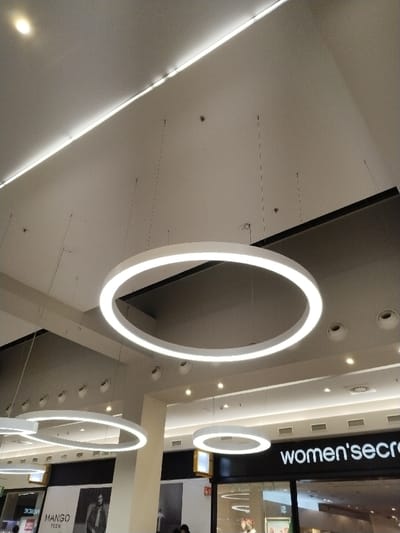 The image showcases the interior of a shopping center with multiple ring-shaped light fixtures suspended from the ceiling. The fixtures are uniformly illuminated, providing ambient lighting. Below, storefronts are visible, including "Mango Teen" and "Women'secret." The ceiling also features recessed lighting and a linear light strip along the top. The architecture is modern, characterized by clean lines and a neutral color palette.