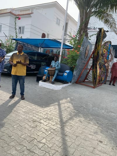 The scene depicts an outdoor paved area in front of the Nike Art Gallery in Ikate, Lagos, Nigeria, under natural daylight conditions.
In the foreground, a light grey interlocking paver surface extends across the ground, featuring distinct shadows from subjects and overhead structures.
On the left, a male subject stands, facing right, wearing a yellow and black patterned polo shirt, blue denim jeans, and dark sandals. His hands are clasped at waist level, appearing to hold a small, dark object.
To his right, a dark-colored sedan, possibly black, is parked beneath a blue fabric shade canopy. Text "WE ARE AHEAD" and "Nike Art Gallery" are visible on or near the vehicle.
Further right, a male subject is seated on a custom-made blue sofa, constructed from repurposed metal barrels, with a dark cushion. This subject wears a blue and white patterned long-sleeved shirt and matching trousers. He is actively playing a djembe drum positioned between his legs.
Behind the seated drummer, two large, vertical art installations are displayed. The left piece features a stylized face and geometric patterns in shades of grey and black. The right piece is a multi-colored, complex sculpture resembling an abstract butterfly or mechanical insect, adorned with various materials and textures.
The background features two large white buildings. The structure on the left has multiple windows and a balcony with pink bougainvillea vines. The building on the right, identified by a sign as "Nike Art Gallery," exhibits textured, wave-like patterns on its facade. A tall palm tree is visible on the far right, alongside various green foliage and plants with orange-red flowers.
Additional subjects are partially visible: one on the far left, wearing patterned attire, and another on the far right, wearing a red/maroon outfit. No direct interactions between the visible subjects are clearly depicted.