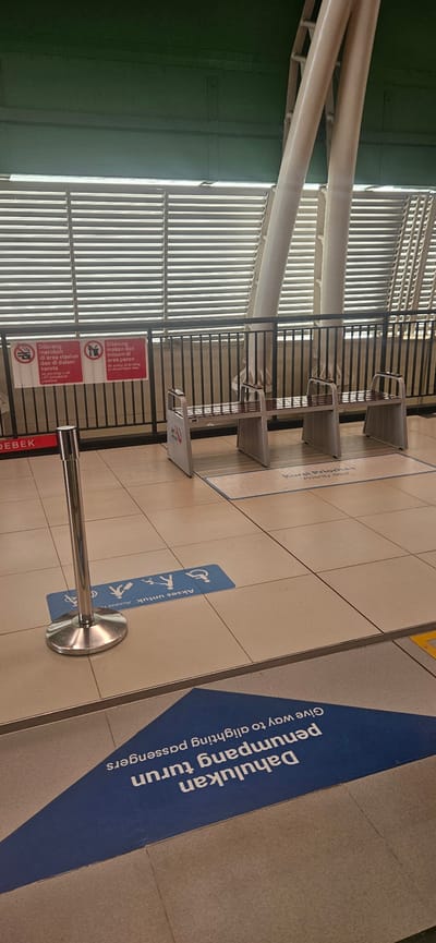 The image shows a platform at a train station in Bekasi, Indonesia. The platform is tiled in a neutral beige color. In the foreground, there is a blue triangular sign on the floor with white text that reads "Dahulukan penumpang turun, Give way to alighting passengers". To the left of this sign is a silver pole with a rounded base. Further back, there are two blue rectangular signs on the floor, each depicting wheelchair and pedestrian icons, with text that reads "Akses Untuk", which translates to "Access for". Two benches with metallic frames and dark seats are visible on the platform. Behind the benches, there is a black metal railing. Above the railing, there is a wall covered with horizontal metal louvers. Two large, light-colored pillars support the ceiling structure. Two red and white signs are attached to the railing; one reads "Dilarang merokok di area stasiun dan di dalam kereta", with "No smoking in all LRT Jabodebek premises" below it. The other sign reads "Dilarang makan dan minum di area peron", with "No eating or drinking in the platform area" below it. The overall environment appears to be an indoor or semi-indoor train station platform.