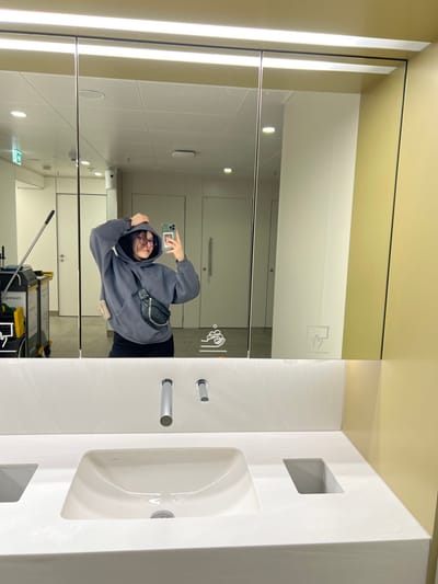 The image displays a modern public washroom facility, stated to be located in Fischamend, Austria. A young adult female is centered in a mirror selfie, captured holding a light-colored smartphone with her right hand. She wears a dark gray hooded sweatshirt with the hood pulled over her head, dark-framed glasses, and black trousers. A black fanny pack with a silver-colored chain is worn diagonally across her chest. Her left hand is raised, subtly adjusting her hood. She exhibits a slight smile.
The washroom environment visible in the reflection features light-colored walls and multiple white closed doors, indicative of stalls or utility rooms. On the far left of the reflection, part of a yellow and black cleaning cart is discernible. The foreground of the image shows a white countertop with an integrated rectangular white sink basin. Two chrome-finished fixtures, a faucet and a sensor (likely for soap or hand dryer), are mounted above the sink. A long, horizontal light fixture illuminates the area above the mirror. Handwashing and hand-drying icons are present on the mirror's surface. A solid beige-yellow wall is visible to the right of the mirror frame.