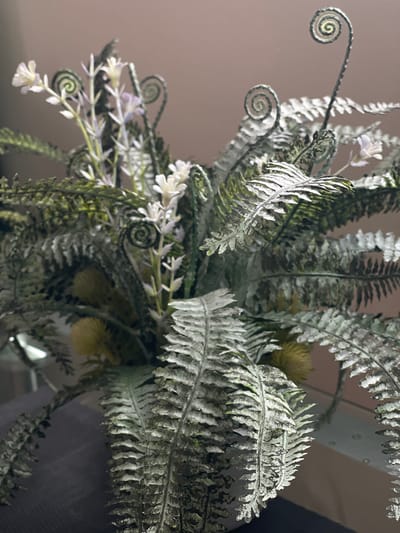 The image shows a close-up of a floral arrangement, primarily composed of artificial ferns and small white flowers. The ferns exhibit a distressed or aged appearance with a dusty, silvery-green hue. Some fern fronds are curled in a spiral shape, characteristic of fiddleheads. Interspersed among the ferns are delicate stems with small white blossoms and a few yellow, spherical elements that could be artificial berries or seed pods. The arrangement is placed in a container, the edge of which is partially visible at the bottom of the frame. The background is a plain, muted color, likely a wall. The lighting suggests an indoor setting, with a soft, diffused light source. There are no people or animals visible in the image. The image's composition focuses on the textures and details of the artificial plants. There is no information in the image to indicate a location in Bolshoy Kamen, Russia.