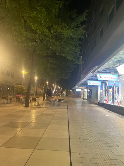 The image depicts a street scene in Pforzheim, Germany, at night. The perspective is from a low angle, looking along a pedestrianized zone. On the right side of the frame is a wide, paved walkway. Rows of tables and chairs, presumably belonging to outdoor cafes or restaurants, are arranged along the edge of the walkway. Trees are planted along the street. The left side of the image shows a commercial building with illuminated storefronts. Several signs for "TUI ReiseCenter" are visible. The street is lit by streetlights. The scene appears quiet, with no people clearly visible.
