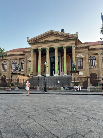 This is a vertical, eye-level shot of a grand building with neoclassical architecture, likely the Teatro Massimo in Palermo, Italy. The building features a prominent portico with tall Corinthian columns and a pediment adorned with decorative carvings. Green lights illuminate some of the columns, creating a striking contrast with the beige stone of the facade. Large arched windows and decorative circular elements are visible on the upper levels.
In front of the building, a paved stone plaza stretches out, and a dark metal fence separates the plaza from the building's entrance area. Several people are scattered throughout the scene. Near the fence, a group of individuals dressed in white are seated, possibly taking a break or waiting. Further to the right, a couple stands in the plaza, with the woman wearing a white dress and the man in a dark shirt. Other individuals are seen sitting or standing on the steps and in the plaza.
The sky is clear and bright, suggesting it is daytime, likely late afternoon given the soft lighting. The overall atmosphere appears relaxed and touristy, with people enjoying the public space in front of the historic landmark. A palm tree is visible on the left side of the frame, indicating a warm climate. Some text is visible on the facade of the building above the columns, but it is too small and blurry to be read.