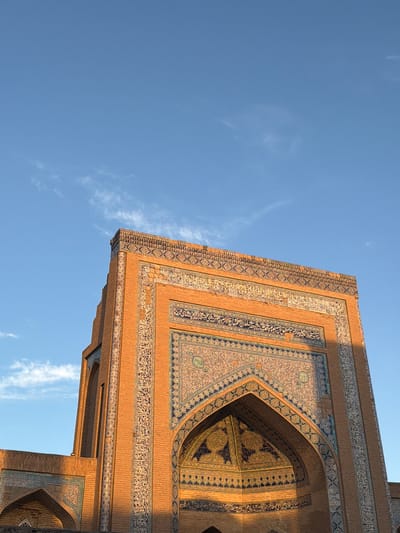 This is a low-angle, vertical shot of a building facade in the city of Khiva, Uzbekistan, likely part of a madrasah or mosque. The building is constructed of brick and adorned with intricate blue and gold tilework, featuring geometric patterns and Arabic calligraphy. The sky above is a clear, bright blue with a few wispy white clouds. The sunlight illuminates the right side of the building, casting shadows and highlighting the textures of the brick and tiles. A portion of an arched entryway with similar tilework is visible on the lower left.