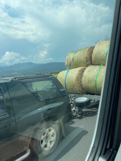 The image presents a roadside scene in Karnare, Bulgaria, captured from the perspective of a vehicle's passenger-side window, indicated by the white frame and black gasket visible on the right. A dark-colored SUV occupies the midground, its rear section, including a roof rack and a silver side step, visible. The SUV's rear tire and wheel appear somewhat soiled. It is actively towing a light-colored utility trailer.
The trailer carries four large, round hay bales, stacked two high and two wide. These bales are secured with two distinct straps: a blue strap vertically positioned over the left stack of bales and a green strap over the right stack. A single spare tire with a silver rim is mounted horizontally on the forward frame of the trailer.
In the background, a range of dark, hazy mountains rises under a sky filled with scattered white and grey cumulus clouds. Between the road and the mountains, expansive green fields and faint indications of a small settlement are discernible. The overall lighting suggests daytime conditions with partial cloud cover.