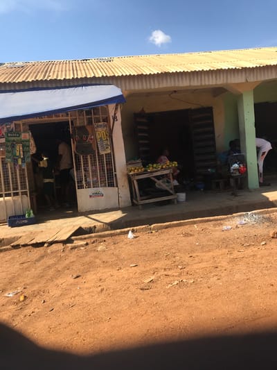 This media file captures a vibrant street scene in Ilorin, Nigeria, under bright daylight. The image is oriented vertically but depicts a horizontal stretch of an unpaved, reddish-brown dirt road next to a multi-story building housing small businesses.

On the left, the building features corrugated metal roofing, possibly on an overhang or upper floor, against a clear blue sky with a solitary white cloud. The building's facade is painted in muted tones, with a light green section on the upper floor and a lighter beige/yellow on the lower levels.

Several individuals are visible. On the upper floor balcony, a person in a white garment is seated. Below, another person is seen near a red and silver generator set, suggesting an off-grid power source. On the ground floor, a vendor is seated behind a wooden stand laden with yellow and green fruits, likely mangoes or oranges. Adjacent to this fruit stand is another shop, partially obscured by a metal gate or security bars. Inside, people are visible, and outside, crates of beverages, including "Delta" brand drinks and a Pepsi crate, are stacked. Various advertisements and signs are affixed to the shop front, with partially legible text including "POS" and "TEL 7033 946" and possibly "SNOW" or "SNOWS" written on the wall. A blue awning extends from one of the shops.

The road surface is dusty and uneven, with scattered small debris and rocks. A prominent dark shadow on the right side of the road suggests strong sunlight from a high angle, indicating it's likely midday. The overall scene portrays daily commerce and life in a bustling urban or semi-urban environment.