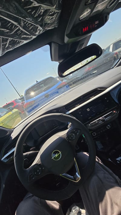 The image is taken from the interior of a car, looking forward through the windshield. The dashboard and steering wheel are visible. The steering wheel has the Opel logo in the center. The car appears to be parked in an outdoor lot with other vehicles visible through the windshield. The sky is clear and blue, suggesting a sunny day. The overall impression is of a new car interior, possibly at a dealership or awaiting delivery. The location is the city of Pedrola, Spain, specifically in an area with a car lot, likely the Opel dealership.