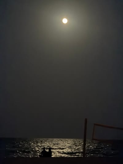 A nighttime scene captures a full moon illuminating the sky and reflecting on the ocean's surface. In the foreground, two silhouetted figures sit on the beach, facing the water. To the right, a volleyball net stands on poles, partially visible against the dark sky. The image appears to be taken from Hollywood Beach, Florida, USA.