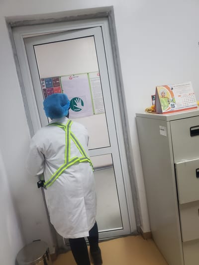 In Gwagwada, Nigeria, a person wearing a white lab coat, a blue hair net, and a reflective vest is standing in front of a glass door. The person is positioned with their back to the camera, facing the door. A green "PUSH" sign with a handprint is visible on the door. To the right of the door, a metal filing cabinet holds a calendar displaying June 2020. The environment appears to be an indoor space with white walls and a yellow floor.
