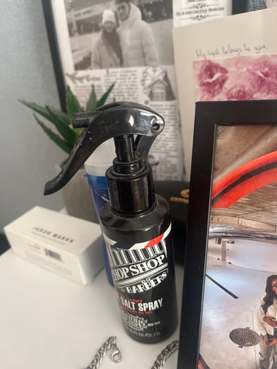 An indoor scene displays a collection of personal items arranged on a light-colored surface, likely a desk or shelf.
In the foreground, a silver-colored curb chain with a lobster clasp rests on the surface. To its left, a white rectangular box with "JASON MARKK" printed on it is partially visible. Centrally, a black plastic spray bottle with a trigger handle stands upright. Its label indicates "BARBERSHOP," "SEA SALT SPRAY," and "NEW YORK." Behind this bottle, a blue and white cup is partially visible.
Further back and to the left, a small green succulent plant in a light-colored pot is present. Behind the plant, a black and white photograph in a light frame shows two individuals in winter attire, smiling. Directly behind this photograph and the plant, a piece of printed material, possibly a newspaper or magazine, displays text, including what appears to be Cyrillic script with "Олександр Невский" partially visible.
To the right of the plant, a white card or note bears handwritten text reading "My heart belongs to you," accompanied by two pink lipstick imprints. The right side of the scene is dominated by a black rectangular picture frame. Inside this frame, a color photograph depicts a segment of an arched, possibly stone or concrete, architectural structure. A prominent red and black striped object arcs through the upper portion of this framed image. In the lower right of the framed photograph, a person, likely a woman with dark hair, wearing a light-colored top and pants, stands on a paved surface next to a manhole cover. An arm is visible around her waist.
