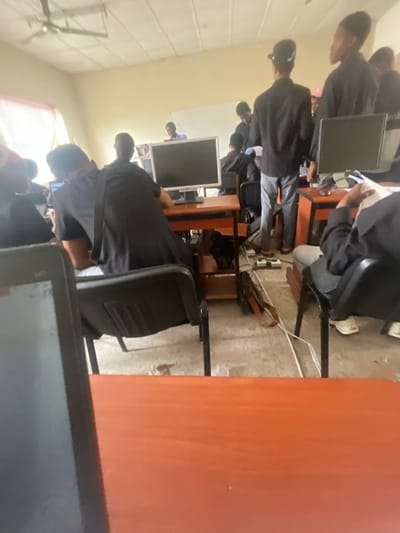 This image depicts an indoor scene within a computer lab or classroom located in Okigwe, Nigeria. Approximately 10-15 individuals, predominantly young adults, are present in various states of activity. Most individuals are wearing black tops or shirts.
Multiple individuals are seated at wooden desks featuring orange tops and dark bases, with several black desktop monitors visible on these desks. A person in the foreground is partially obscured by a laptop resting on an orange desk. Another individual, wearing a black shirt and white pants, is seated with their back to the camera at a desk with a monitor. On the right, a seated person holds several white papers.
Standing individuals are positioned throughout the room; one person in a black shirt and grey pants stands in the center-midground with their back to the camera. In the background, near a white rectangular whiteboard mounted on the wall, a person in a blue shirt is visible.
The room features light-colored walls and a light grey concrete or tiled floor exhibiting visible wear and scattered debris. A white four-bladed ceiling fan is centrally mounted on the ceiling. Numerous electrical cables and white power strips are distributed across the floor, connecting to the computer equipment. A long, dark brown object, possibly a carrying case, rests on the floor amidst the cables. The room is illuminated by natural light entering from a window with light-colored curtains on the left and by overhead fluorescent light fixtures.