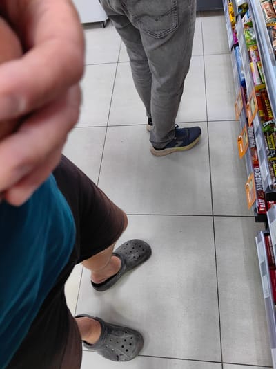 The image shows the legs and feet of two people standing on a tiled floor in what appears to be a retail store. The person in the foreground is wearing dark shorts and dark gray Crocs. The person in the background is wearing gray jeans and blue athletic shoes. A shelf stocked with candy bars and other packaged goods is visible to the right. The environment suggests a consumer setting like a grocery store or a convenience store. The presence of specific merchandise and signage would be needed to confirm the exact location within Myadzyel, Belarus.