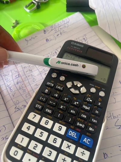 The scene is set in Epe, Nigeria. The image displays an overhead close-up of a desk surface featuring mathematical calculations, a scientific calculator, and a pen held by a hand. A light-skinned hand with a visible fingernail holds a white pen horizontally. The pen has a green band near its tip and is branded "Monica.cash" with a small "MYCC" logo visible on its body. This pen is positioned across the upper section of a black Casio fx-991EX ClassWiz scientific calculator. The calculator's display shows "7-12" on the top line and "=-5" on the bottom line. Various calculator buttons, including numerical keys (0-9), operational symbols (+, -, x, ÷), and scientific functions (sin, cos, tan, log, alpha, shift, optn, calc, solve), are clearly visible. Beneath the calculator and pen, a sheet of white lined paper from a notebook contains handwritten mathematical notes, including formulas (e.g., "b = Σ(x-x̄)(y-ȳ) / Σ(x-x̄)²" and "a = ȳ + b x̄"), and a table with columns for "x", "y", and "x-x̄" containing numerical entries such as "50, 60, 65, 70, 80" and "-4, -2, 0, 2, 4". In the upper left background, a blurred bright green surface is visible, along with a metallic object resembling a key ring component. A crumpled piece of white paper is in the upper right background.