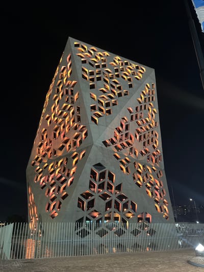 The image shows the Caraffa Fine Arts Museum in Cordoba, Argentina, at night. The museum's distinctive geometric architecture is prominently featured, illuminated with orange light emanating from the interior spaces. The building appears to be constructed of grey concrete with geometric cutouts. Some other architectural elements such as a column are visible on the right. The background is predominantly dark.