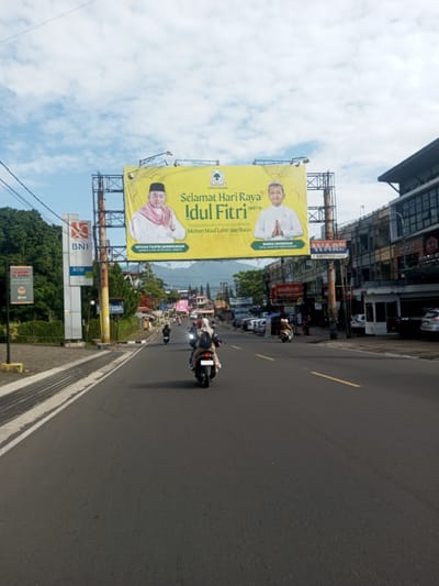 The image shows a street scene in what appears to be an urban area, possibly in Southeast Asia. The main focus is a large yellow billboard spanning the road, displaying a greeting for "Selamat Hari Raya Idul Fitri" (Happy Eid al-Fitr) along with portraits of two men and the logos of political parties.
Below the billboard, a road with a yellow center line stretches into the distance. Several motorcycles are traveling on the road, with one in the foreground carrying two individuals.
On the left side of the road, there is a BNI (Bank Negara Indonesia) ATM and signage. On the right side, various commercial buildings are visible, including stores like "erafone" and "sorio."
The sky above is partly cloudy. The overall scene suggests a bustling commercial district during a festive season. Based on the road signage and language on the billboard, the location is most likely in Indonesia rather than Myanmar.