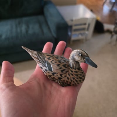A hand holds a carved wooden duck in Portland, Oregon. The duck is brown and black with detailed feather patterns and a dark bill. The hand appears to be an adult's, with visible lines and a pinkish hue. In the background, out of focus, is a dark green couch and a blurred white crib-like structure. The flooring appears to be a light-colored carpet or wood.