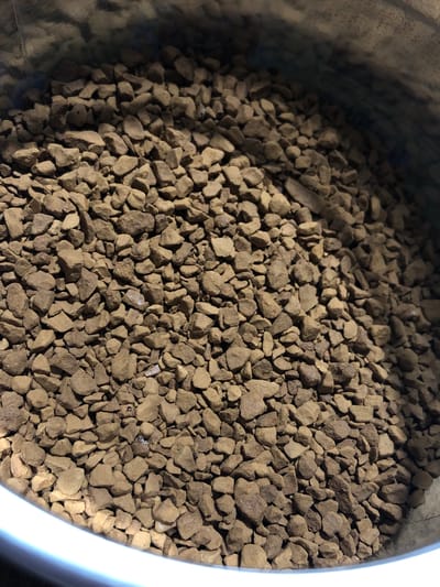 This media file is a close-up, top-down shot of instant coffee granules filling a metallic container. The granules are small, irregular in shape, and a uniform brown color, with some lighter and darker variations suggesting variations in roasting or particle size. The container's rim is visible on the right and left edges, appearing as a smooth, curved metallic surface. The lighting creates shadows, particularly on the left side, indicating the light source is likely coming from the right. There is no visible text in the image. The scene is likely indoors, given the controlled lighting and focus on the product. No people or animals are present. The image focuses solely on the texture and composition of the coffee granules. The location context of Yelizovo, Russia, does not provide any specific visual cues within the image itself.