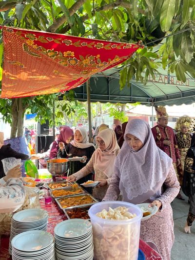 A wedding reception is being held outdoors in Kulam Baro, Indonesia, under a canopy of trees and a green tent. A red and gold patterned cloth is draped overhead. Several women in modest Islamic attire, including hijabs, are serving and receiving food from a buffet table. The buffet table is laden with various dishes in metal trays, including what appears to be curry, noodles, and fried items. Stacks of white plates and bowls are arranged on the table. In the background, a man and woman dressed in traditional wedding attire, with elaborate gold headwear, are visible. The atmosphere appears festive and communal, typical of a local celebration.