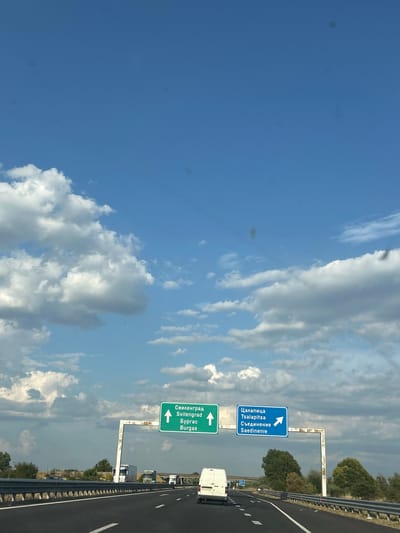 The image depicts a highway scene in Malo Konare, Bulgaria, under a partly cloudy blue sky. The perspective is from the driver's seat of a vehicle traveling on a multi-lane asphalt road. Two overhead green and blue road signs provide directional information. The green sign indicates "Свиленград / Svilengrad" and "Бургас / Burgas" with an upward arrow, suggesting these destinations are straight ahead. The blue sign indicates "Цапарица / Tsalapitsa" and "Съединение / Saedinenie" with a rightward arrow, implying a turn is needed for these locations. Several vehicles are visible on the highway, including trucks and white vans. The road is bordered by a metal guardrail and an embankment with vegetation and trees. The overall environment suggests a sunny day on a major roadway.