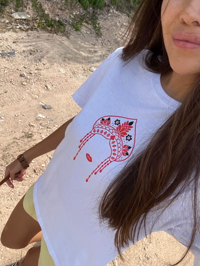An overhead medium shot captures a woman wearing a white t-shirt with a red graphic of a face and yellow shorts. The graphic on her t-shirt features red lips and an elaborate headdress with floral and geometric patterns in red, black, and white. The woman is holding a brown leash in her left hand, suggesting she might be with a dog, although the dog is not visible in the frame. Her long, dark brown hair partially obscures the t-shirt and her right shoulder. She is standing on a path composed of dirt and small stones, indicative of an outdoor, possibly rural or park-like setting. The background consists of more dirt, scattered rocks, and some green foliage, hinting at a natural environment. The lighting suggests it is daytime, with sunlight casting shadows on the ground. The overall scene appears to be a candid shot, possibly taken while walking. The specific location in Baan Nok Sai, Thailand is not discernible from the visual information provided.
