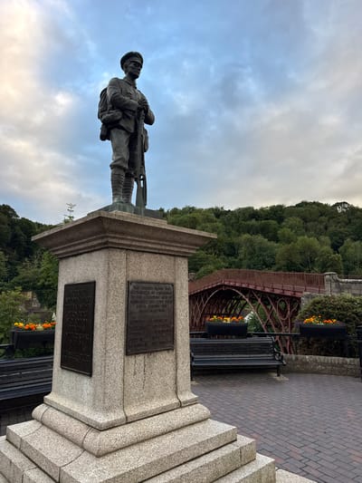 The image shows a war memorial in Ironbridge, United Kingdom. The memorial features a bronze statue of a soldier standing at attention, holding a rifle. The statue is mounted on a granite pedestal with two bronze plaques. The left plaque reads "In grateful and undying memory of the valiant men of Ironbridge who laid down their lives in the Great War 1914-1918." The right plaque reads "The church clock was illuminated as a memorial to the following men of Ironbridge who lost their lives during the 1939-1945 War." Below this are a list of names.

In the background, there is a red wrought-iron bridge, a wooded hillside, and a cloudy sky. Benches and flower planters are visible in the foreground, suggesting a public space or park. The scene appears to be captured during daylight hours, with overcast conditions.