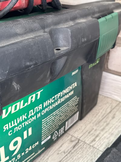 The image shows a close-up, low-angle shot of a black and green plastic toolbox, likely on a wooden floor. The toolbox is dusty. Text on the green section of the toolbox is visible, partially obscured. The visible text includes "VOLAT", "ЯЩИК ДЛЯ ИНСТРУМЕНТА", "С ЛОТКОМ И ОРГАНАЙЗЕРАМИ", a large "19", and dimensions "77,5 x 24 cm". There is also a barcode and the "EAC" certification mark. The red and black cord of an electrical tool is visible entering the top of the toolbox from the left. The environment appears to be indoors, possibly a storage area or workshop, in the city of Yarominski Sel'skiy Sovet, Belarus.