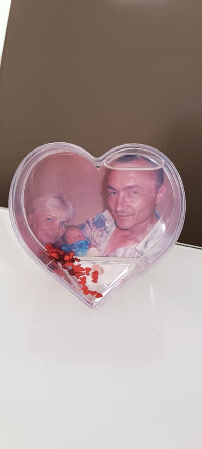 The image displays a heart-shaped photo frame containing a photograph of three people. In the photograph, a woman with blonde hair is on the left, a baby in blue is in the center, and a man with dark hair is on the right. The man is wearing a patterned shirt. The photo frame is made of clear plastic and contains red heart-shaped confetti. The frame itself is resting on a white surface. The background is a solid brown color, suggesting an indoor setting. The lighting appears to be artificial, possibly from a lamp or overhead lighting. There is no visible text in the image. The overall scene evokes a sense of family and affection, with the heart shape and confetti emphasizing love and celebration. The image was taken in Winnenden, Germany.