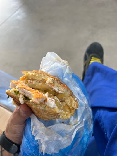 A high-angle, close-up shot captures a person's hand holding a large, cut sandwich, likely a street food item, in Sevlievo, Bulgaria. The sandwich is densely packed, revealing its contents: golden-brown fried chicken pieces, a generous amount of white creamy sauce (resembling mayonnaise or a similar dressing), French fries, and small hints of green leafy elements like herbs or lettuce. The bread appears to be a toasted bun or thick flatbread. It is wrapped in a translucent white paper or wax paper, which is further enclosed in a vibrant blue plastic bag.
The hand holding the sandwich is visible in the lower right of the frame, wearing a watch with a dark gray or black fabric strap and a metallic silver buckle. In the blurry background, partially visible in the upper left, is a person's leg clad in bright blue trousers or scrubs. A black shoe with distinctive yellow laces or accents is also visible on the foot. The ground beneath is a uniform light gray, textured surface, consistent with concrete or pavement, suggesting an outdoor or casual setting. The bright, even lighting indicates daytime. There is no visible text in the image.