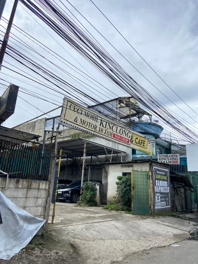 The image shows a street scene in Kecamatan Margahayu, Indonesia (latitude: -6.975, longitude: 107.571). It depicts a small business called "Cuci Mobil Kinclong & Motor 18 Jam Non Stop & Cafe" situated on the right side of the image. The business sign is prominently displayed above the entrance, which is partially obscured by a green metal gate. A black SUV is parked in front of the shop, and there is a sign that says "Vape Store Bandung Vaporizer" next to the gate. There is a white sheet on the ground to the left of the entrance and a significant amount of electrical wiring overhead. The sidewalk and the street are made of concrete and appear to be in good condition. The sky is overcast and there is a hint of rain in the air. There are no people present in the image.