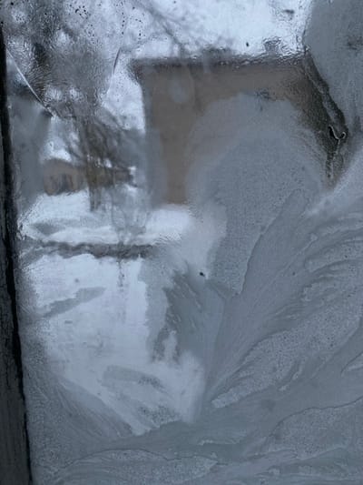 The image shows a frosted window with a view of the outside obscured by ice patterns. Shapes of buildings and trees are visible through the ice. The frost creates intricate textures, with crystalline structures spreading across the glass. The bottom edge of the window is visible with black cracks. The overall tone is cold and monochrome.