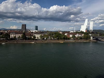 The image captures a panoramic view of the Rhine River in Basel, Switzerland, under a partly cloudy sky. The river dominates the foreground, appearing dark and textured with ripples. Along the far bank, a promenade is visible, lined with trees and buildings. A significant number of people are gathered on the promenade and on steps leading down to the river, suggesting a recreational area. Several boats, including a larger white vessel, are moored or moving on the water.
The cityscape behind the riverbank features a mix of architectural styles. Traditional European buildings with tiled roofs stand alongside modern skyscrapers. Notable are two distinctive, tall white buildings with angular designs on the right side of the frame, and a dark, rectangular skyscraper to the left, creating a contrast between old and new urban development. A bridge crosses the river on the far right. The overall scene depicts a lively urban environment with people enjoying a sunny day by the river.