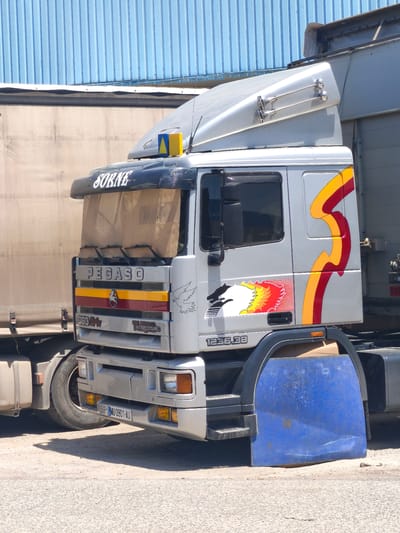 This is a medium shot of a silver Pegaso 300/24 Intercooling semi-truck parked outdoors in Quart de Poblet, Spain. The truck is silver with red and yellow flame-like decals on the side and a black visor above the windshield with the word "SORNE" in white lettering. A light brown shade hangs behind the windshield. Two yellow and blue triangles are mounted on the top of the truck's cabin. The truck has a license plate that reads "SP MU 0901 AU". In the background, there is a light tan trailer and a blue ribbed metal structure, possibly a building or another trailer. The ground is a dirt and asphalt mixture. A blue plastic mud flap is attached to the rear wheel well of the truck.