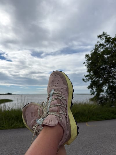 The image shows a close-up of a person's feet wearing light pink and yellow New Balance running shoes, with the shoes positioned at an angle. The person is likely engaged in an outdoor activity, possibly running or walking. The background features a body of water, possibly the sea, with a distant shoreline and trees. The sky is cloudy, with patches of blue visible, suggesting it might be daytime with variable weather. The ground appears to be a paved path, possibly a promenade or a road, with green grass and vegetation lining the side. The overall scene evokes a sense of outdoor activity and a serene, natural environment. Visible text on the shoe includes "New Balance" and "OAE". The location is Haapsalu, Estonia.